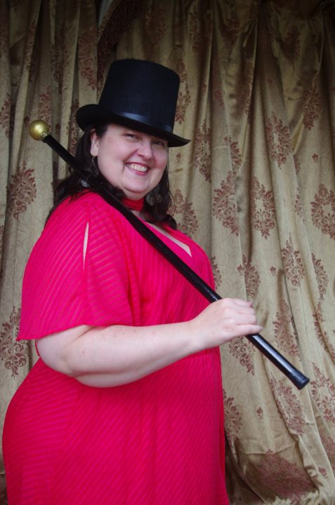 Director Kayt in a bright red dress with wearing a top hat and cane